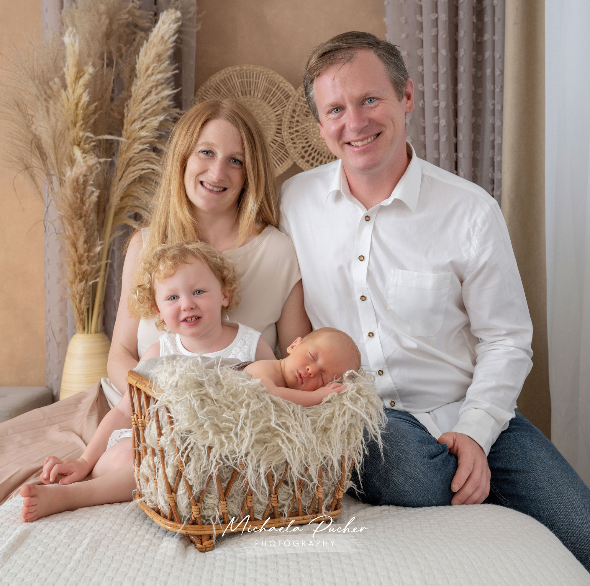 Junge Familie sitzt eng beisammen auf dem Bett; das ältere Geschwisterkind sitzt vorne, während das Neugeborene im kleinen Korb schläft. Eltern lächeln in die Kamera, umgeben von warmen Boho-Elementen.